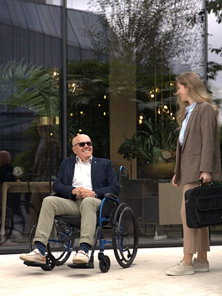 Man seated in a Strongback Comfort wheelchair outside a cafe, talking with a colleague